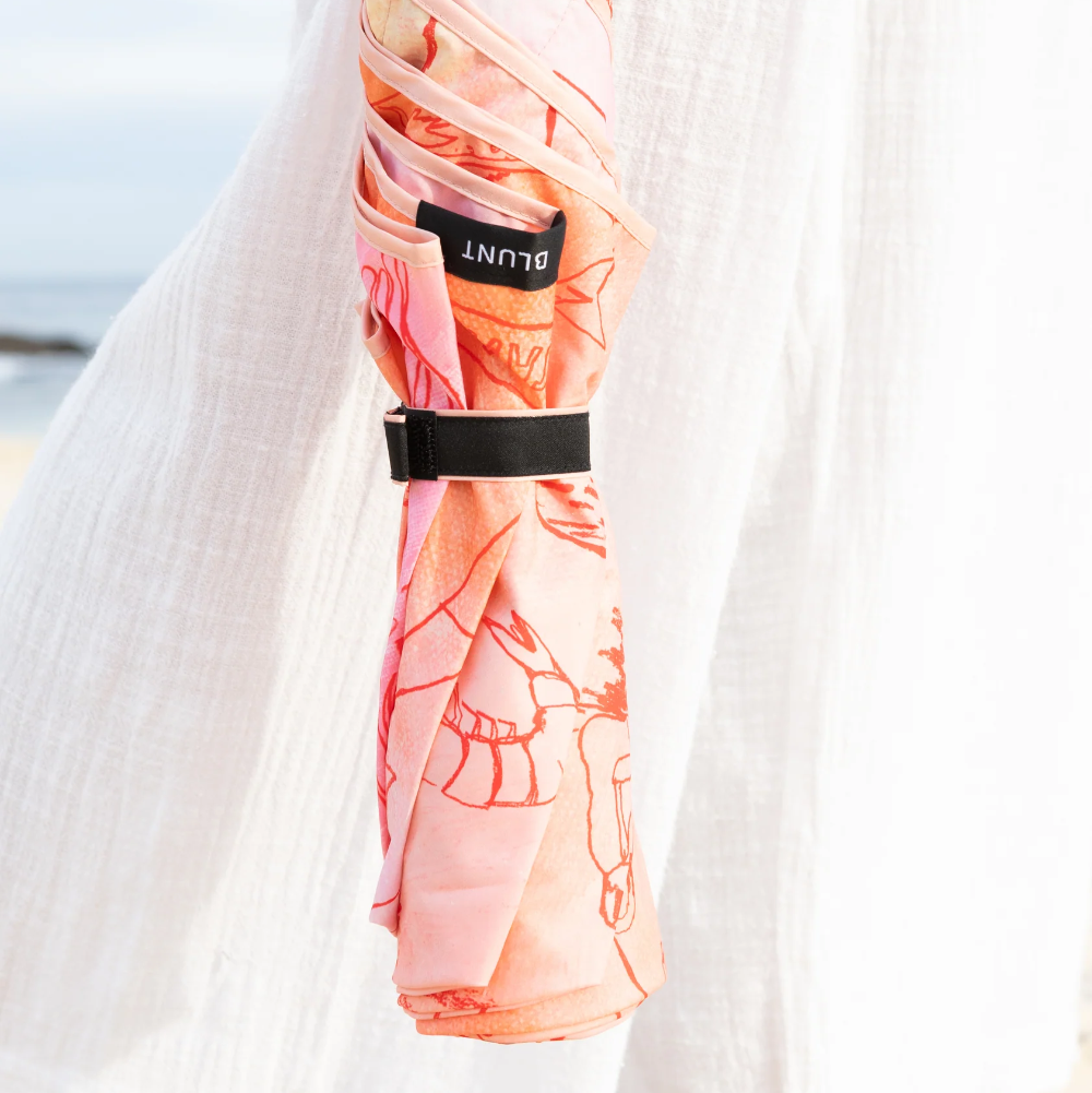 A compact umbrella with a pink and orange floral pattern is wrapped in a white fabric sleeve and held by a person wearing white clothing on a beach.