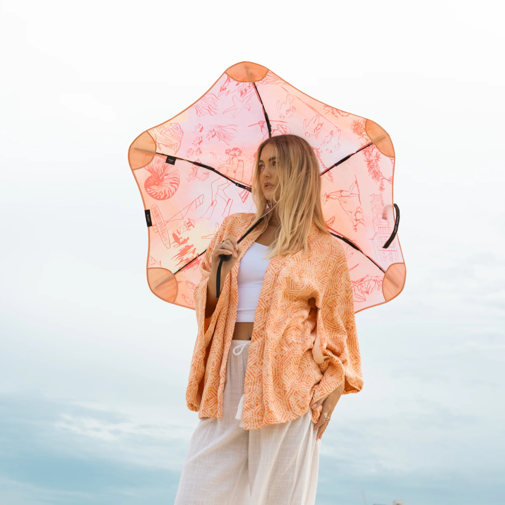 A woman stands outdoors holding a translucent peach umbrella with a floral print. She is wearing a matching peach lace cardigan over a white top and white pants, set against a bright, overcast sky.