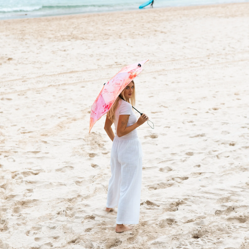 A woman in a white outfit stands on a sandy beach holding a pink umbrella, with the ocean and distant buildings in the background.