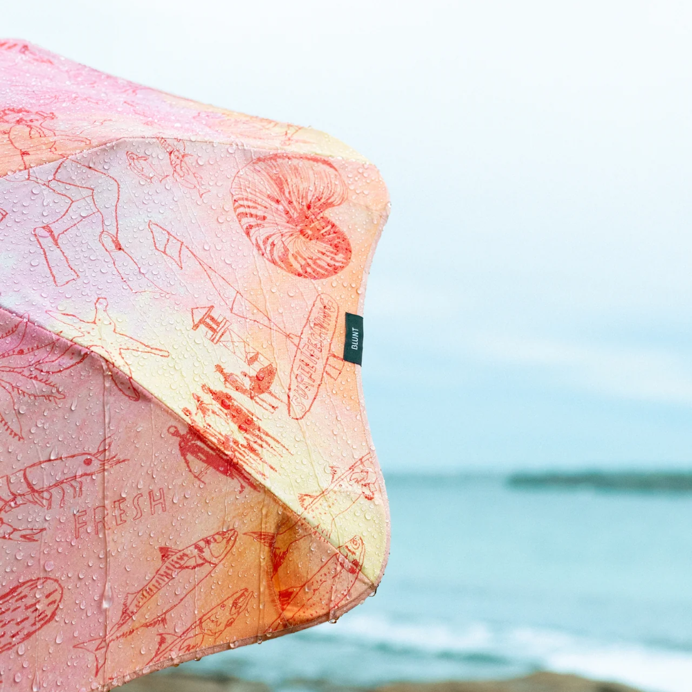 A pink and orange patterned beach umbrella with sea creature illustrations, shown with its canopy open against a blurred coastal background of water and rocks.