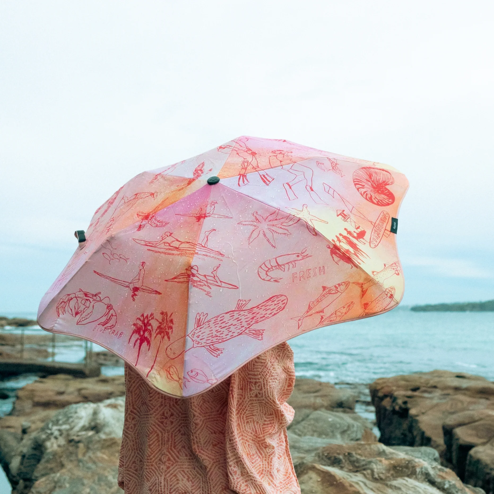 A person standing on rocky shoreline holding a translucent pink umbrella with red tropical fish and shell illustrations, with water and distant land in the background.