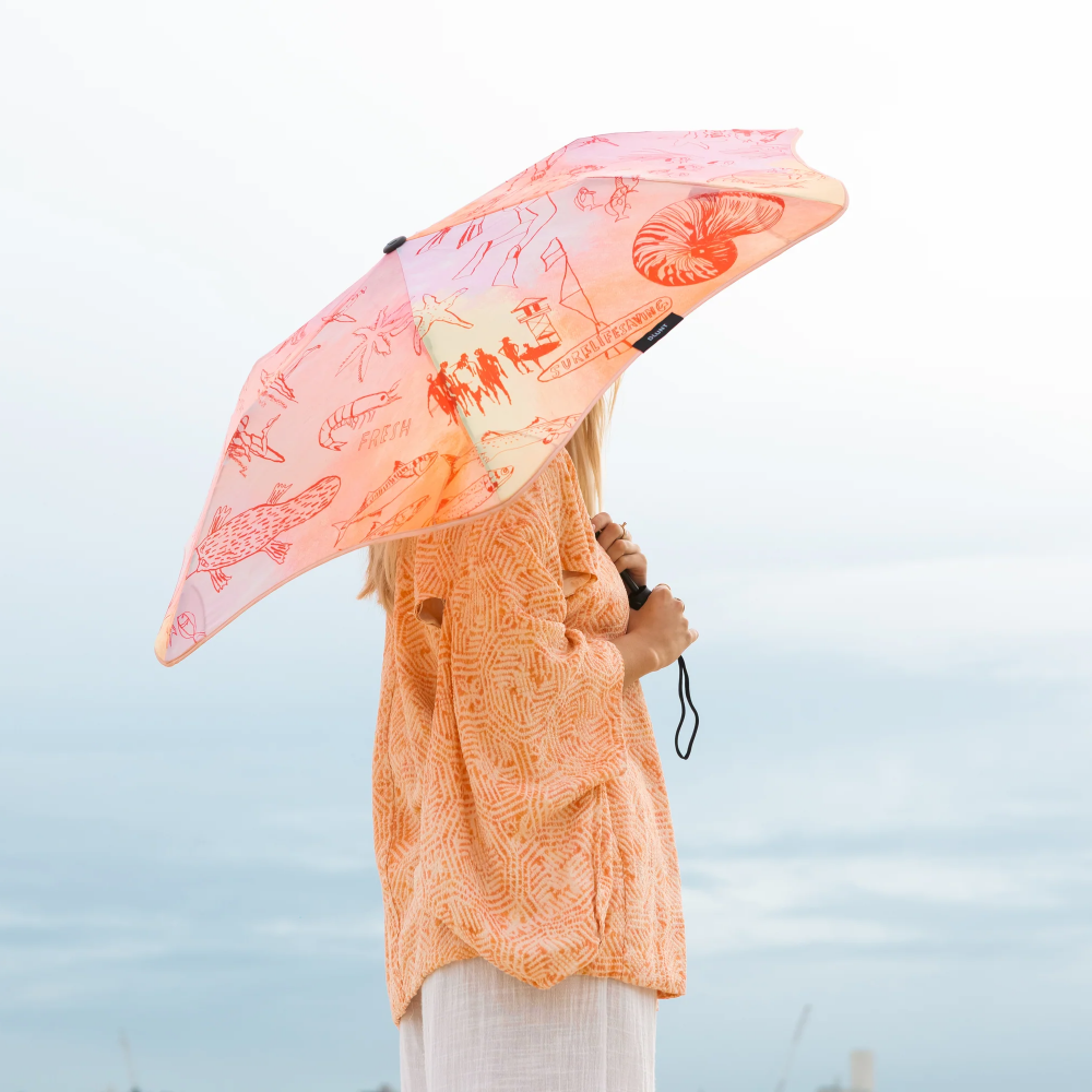 A woman in an orange patterned top and white pants holds a pink umbrella with red sea-creature illustrations, photographed against a pale sky.