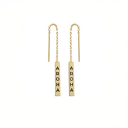 A pair of gold barbell drop earrings with short chains, each featuring a vertical rectangular bar engraved with the word 'AROMA' in black capital letters, shown on a plain white background.