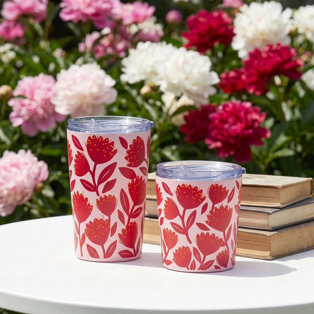 Two tall insulated tumblers with clear lids sit on a white table outdoors, decorated with a red floral pattern on a white background. Books and a blurred garden of pink, red, and white flowers are in the background.