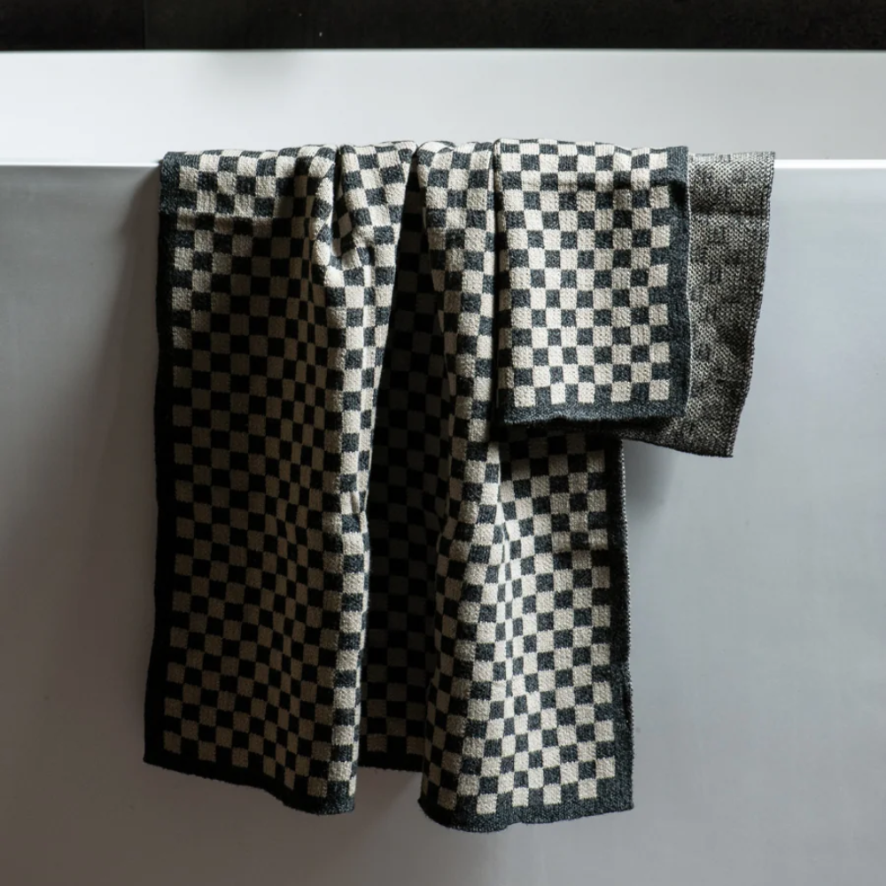 A black and white checkered kitchen towel draped over the edge of a white sink, with a matching folded towel and a dark textured wall with silver faucet handles in the background.