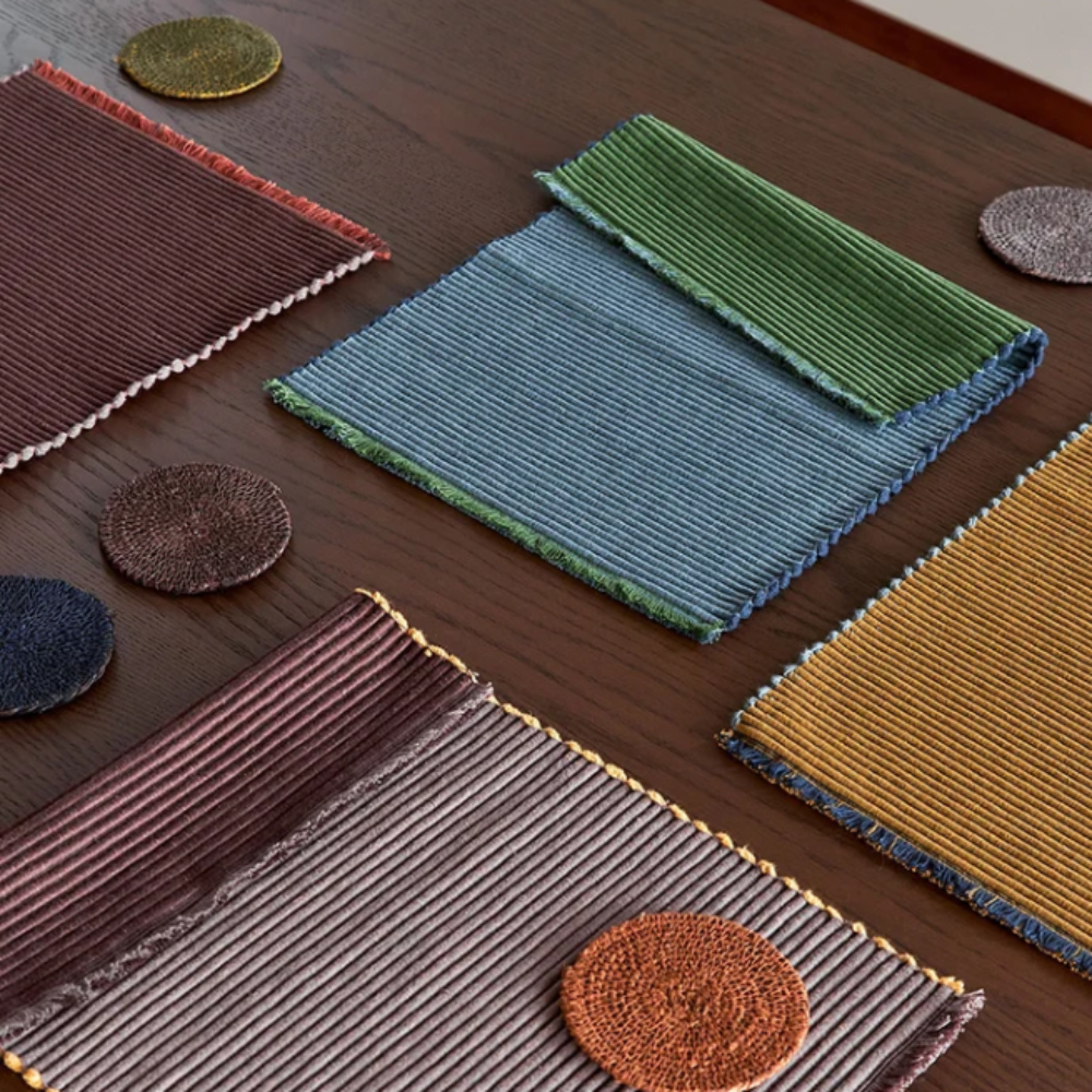 A styled flat-lay of multiple textured woven placemats and coasters in assorted colors, including blue, green, yellow, brown, and gray, arranged on a dark wooden table.
