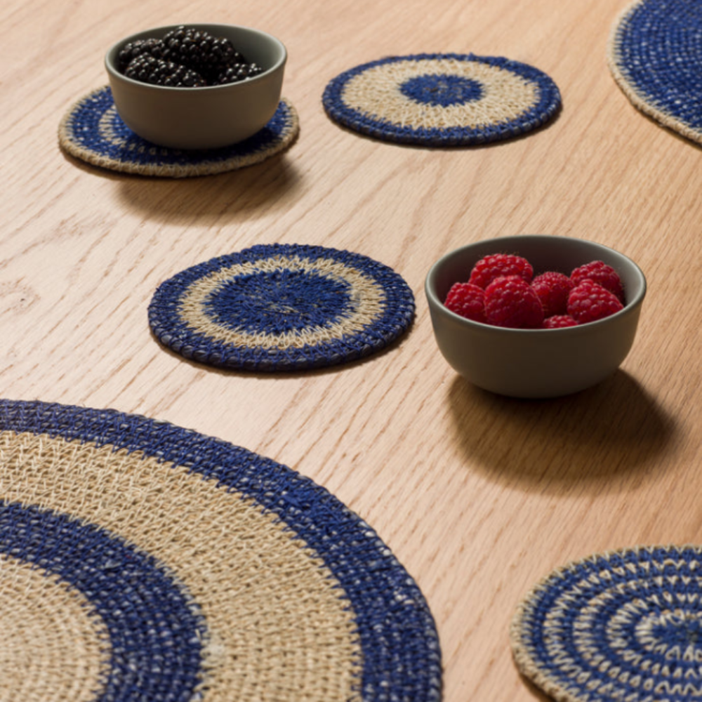 A set of round braided placemats and coasters in blue and beige, styled on a light wood table with two small bowls containing berries.