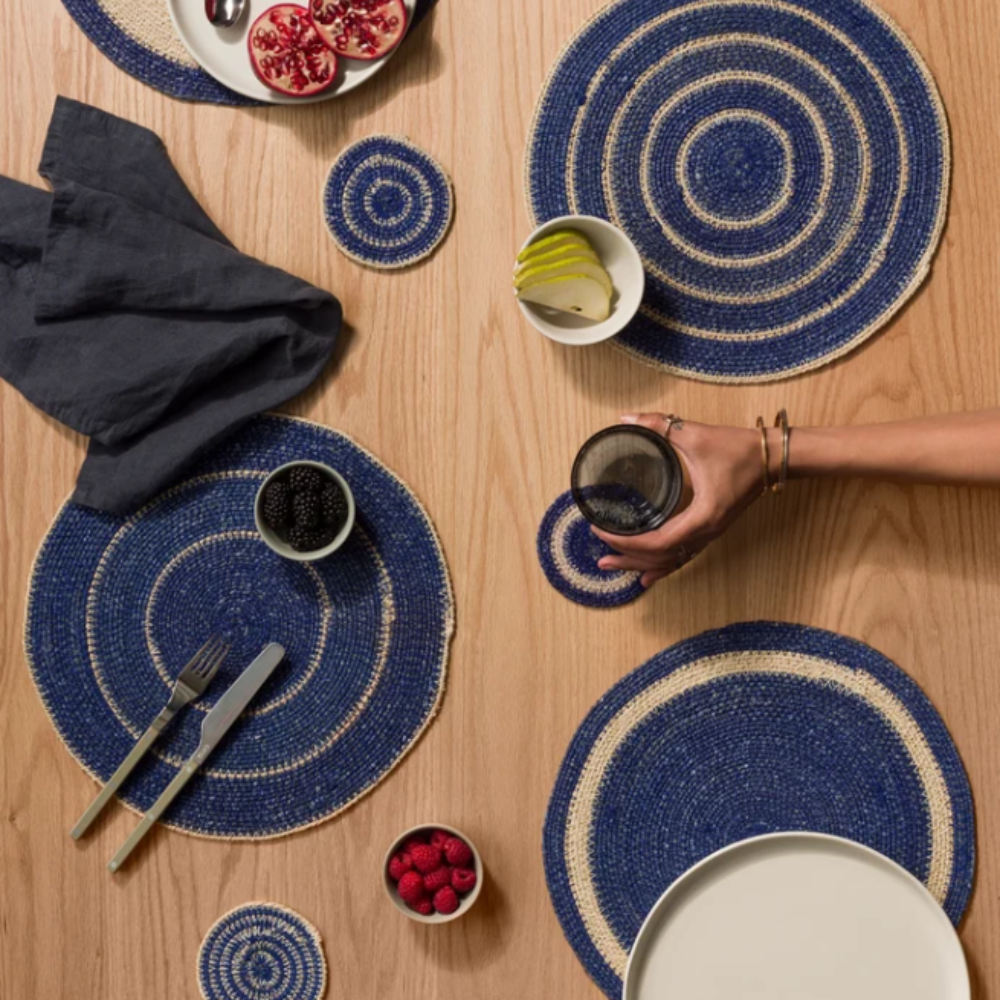 A styled overhead tabletop scene showing multiple round woven placemats and matching coasters in a blue and cream spiral pattern on a light wood table. Plates, cutlery, a glass, fruit, berries, and a hand holding a drink are arranged around the surface, indicating a real dining environment and making this a Lifestyle image.