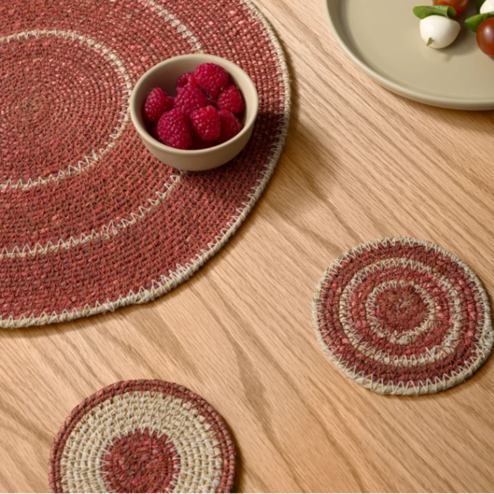 Round woven placemats and coasters on a wooden table with a bowl of raspberries and a plate of salad.