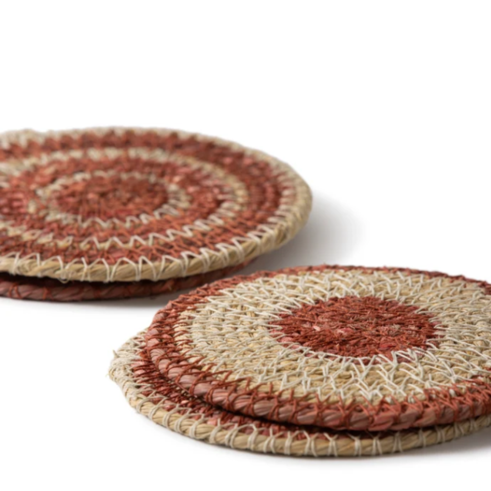 Two round woven coasters with concentric circular patterns in natural beige, rust red, and brown, arranged on a clean white background.
