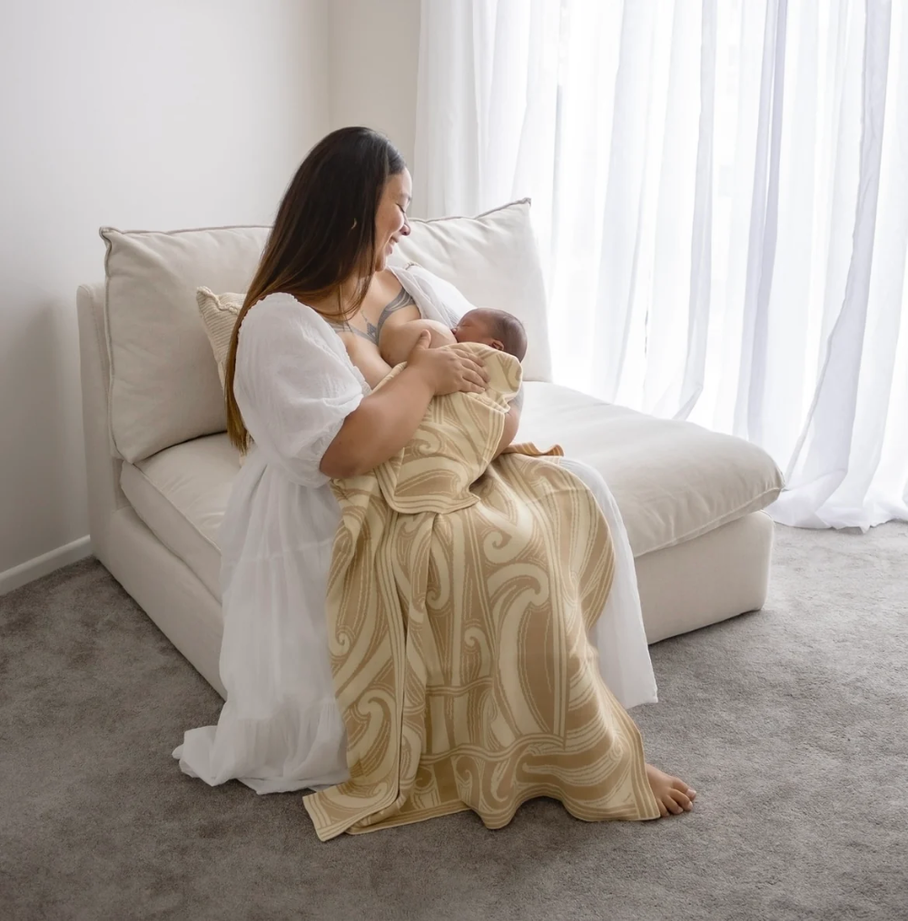 A woman sits on a cream chaise lounge holding a baby wrapped in a beige and tan patterned blanket, in a bright room with white curtains and gray carpet.