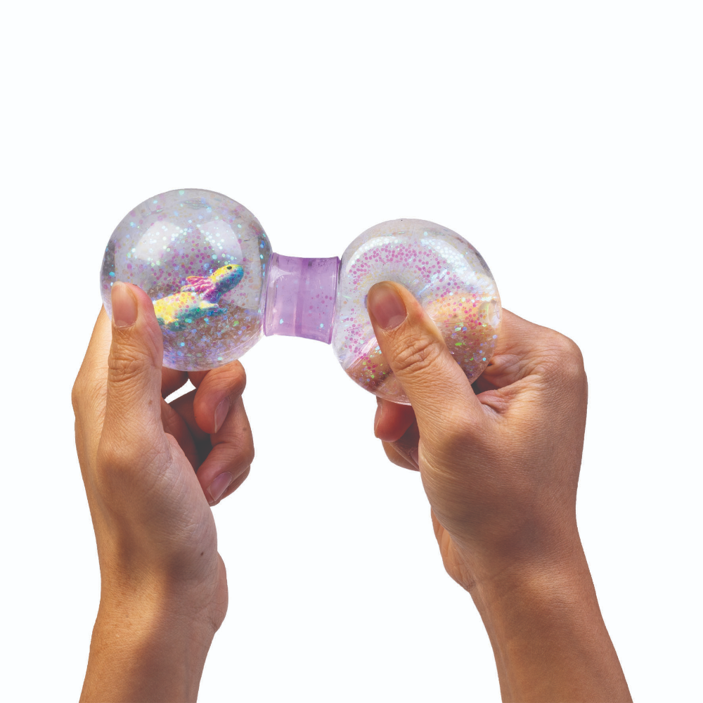 Two hands holding a clear, dual-blob sensory toy with glitter and a small rainbow-colored figure inside, against a solid white background.