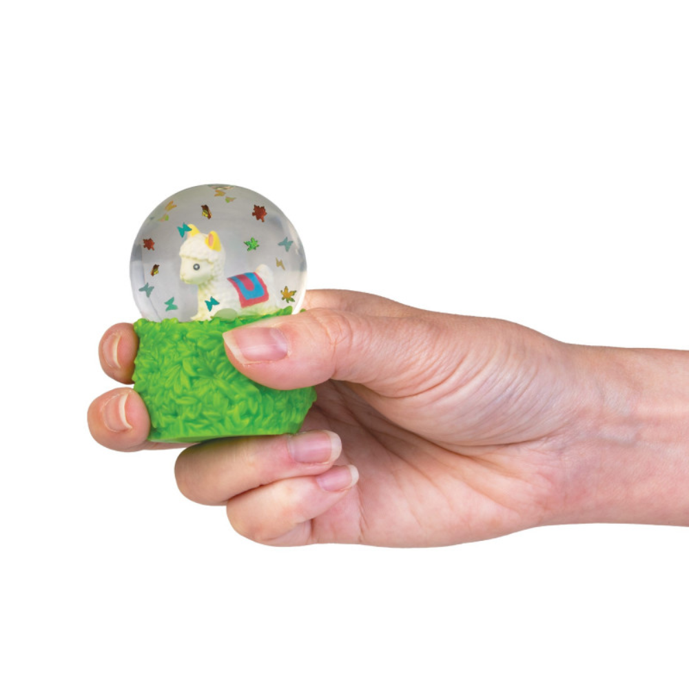 A hand holding a small stress toy snow globe with a green grass-textured base and a clear round top containing a cartoon figure and colorful star details, shown against a plain white background.