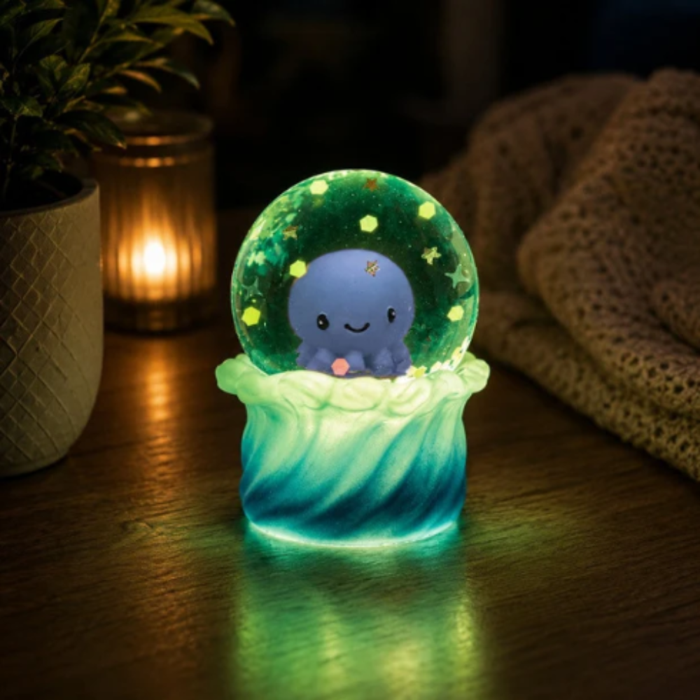 A glowing octopus snow globe night light sits on a wooden table in a cozy, dimly lit room. The octopus is blue and smiling inside a green glittery globe, set on a wavy, light-up base that glows with green and blue light.