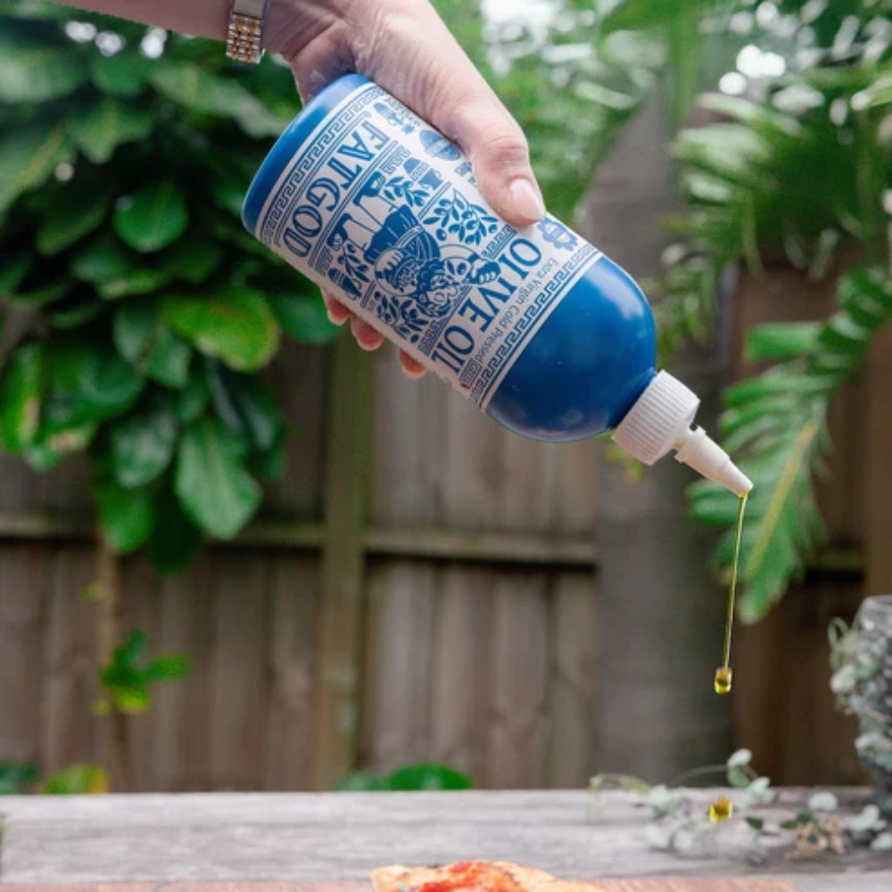 A hand holds a blue squeeze bottle labeled 'PINECONE STREET' while drizzling oil onto three slices of pizza on a wooden board, with a garden fence and green plants in the background.