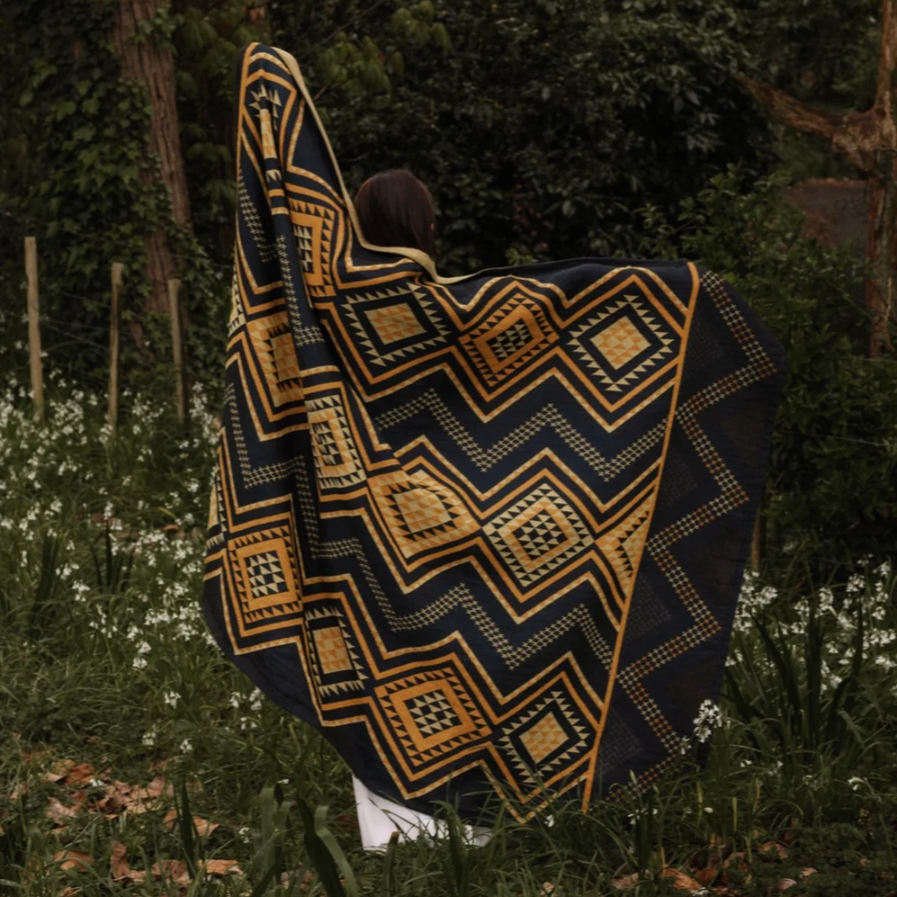 A person standing outdoors in a wooded area with greenery and small white flowers, wrapped in a large patterned scarf or shawl. The textile has a bold geometric design in mustard yellow, navy, and black.