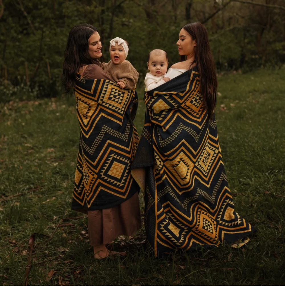 Two women holding babies wrapped in patterned cloths in a forest setting