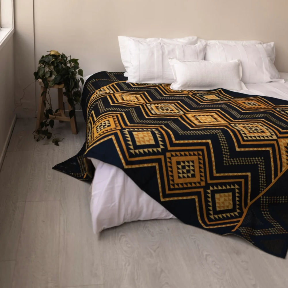 A bed in a minimalist bedroom setting, styled with a dark navy blue and mustard yellow geometric patterned blanket, white pillows, and a small potted plant on a wooden stand next to a window.