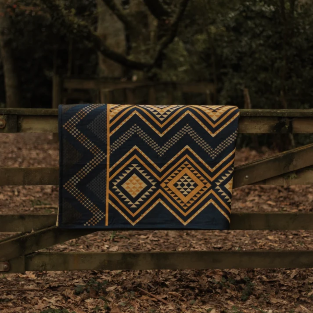 A folded patterned blanket or throw with geometric Southwestern-style motifs in navy blue, mustard yellow, and beige, draped over a rustic wooden fence in a wooded outdoor setting.