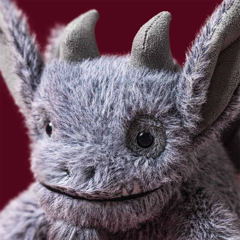 A close-up of a gray furry monster or dragon toy with pointed ears, dark eyes, and a stitched smile against a dark red background.