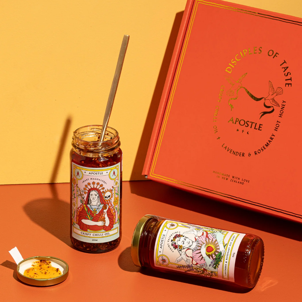 A styled product shot of a spice blend set featuring two glass jars of 'Apostle' spices with illustrated labels, one standing upright and one lying down, a small open lid with yellow spice powder, and a large red-orange gift box on a warm orange and yellow background.