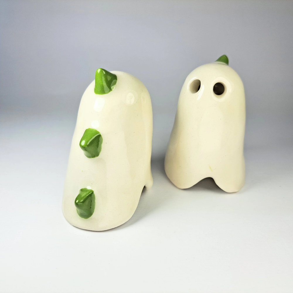 Two ceramic ghost-shaped salt and pepper shakers in off-white with glossy green accents, photographed on a light gray studio background.