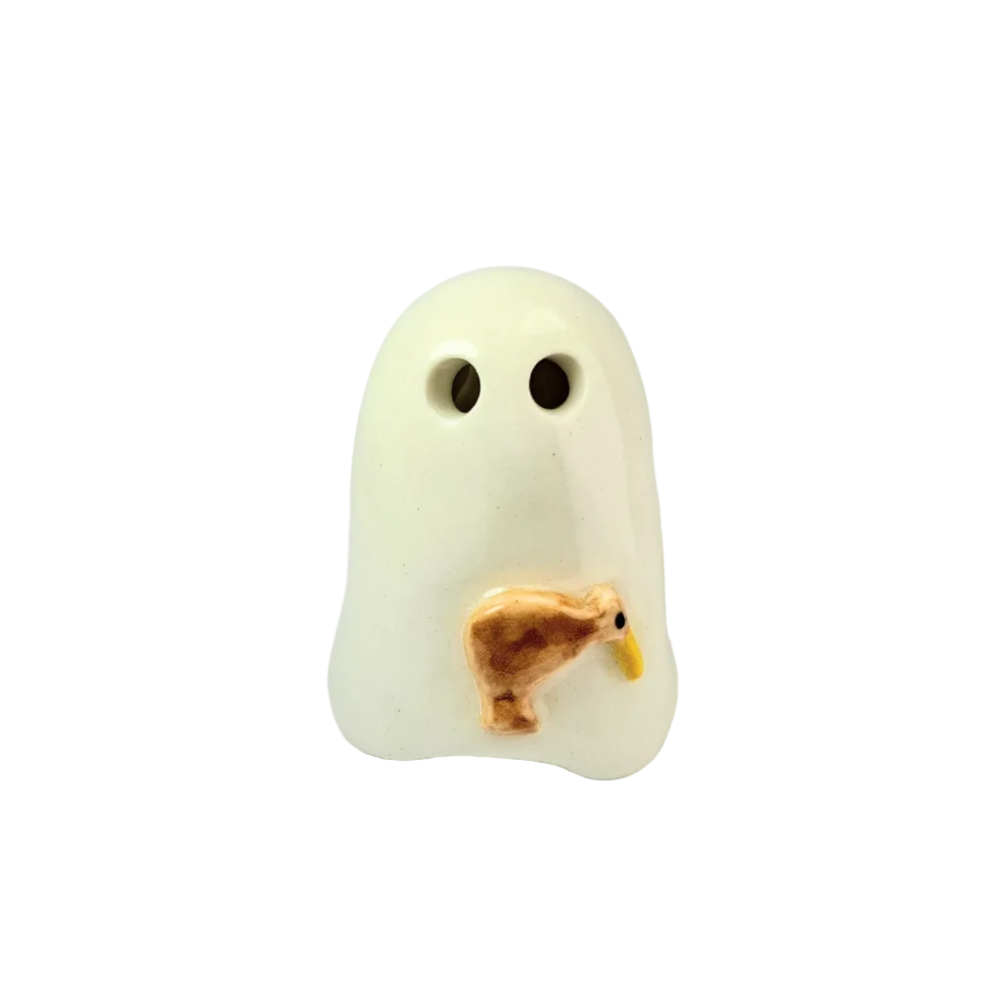 A ceramic ghost-shaped object in warm off-white with two dark eye holes and a small brown kiwi bird figure with a yellow beak attached near the lower front, photographed on a light gray studio background.