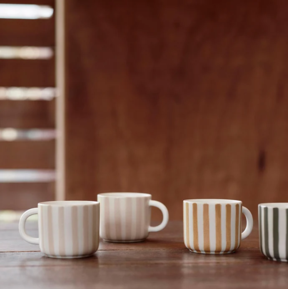 Four ceramic mugs with vertical stripes in neutral and earthy tones, arranged on a wooden table against a warm wooden background.