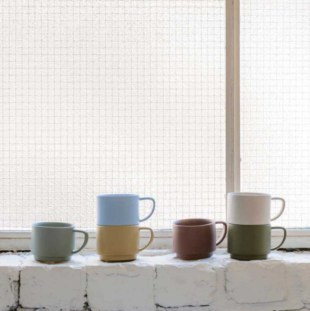 Four stacked ceramic mugs in muted pastel and earthy colors sit on a white brick windowsill in front of a bright window.