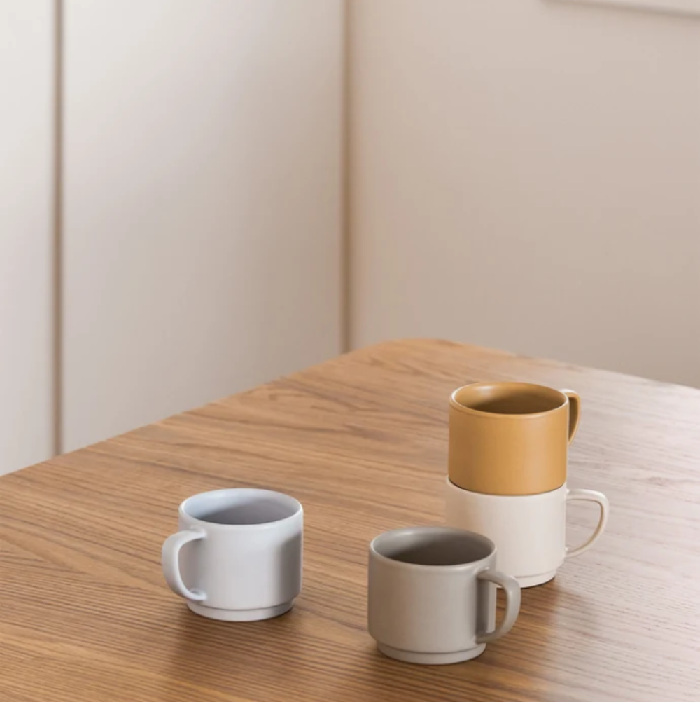 Four minimalist ceramic mugs in neutral tones arranged on a wooden table, with one mug stacked on top of another in the back.