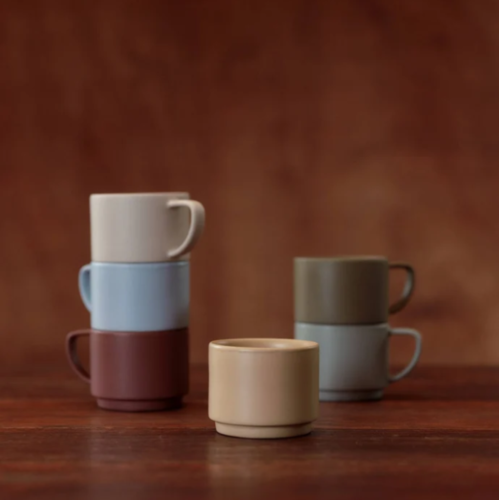 Six small ceramic cups in muted neutral and pastel colors arranged on a wooden surface against a warm brown background. Two stacks are on the left, one stack on the right, and one cup stands alone in front.