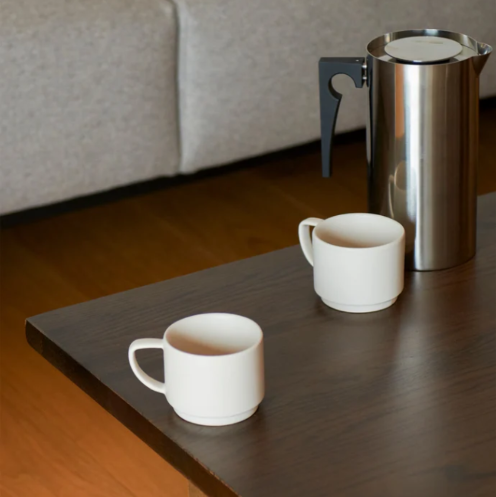 Two white mugs and a stainless steel pitcher on a wooden surface with a neutral background.