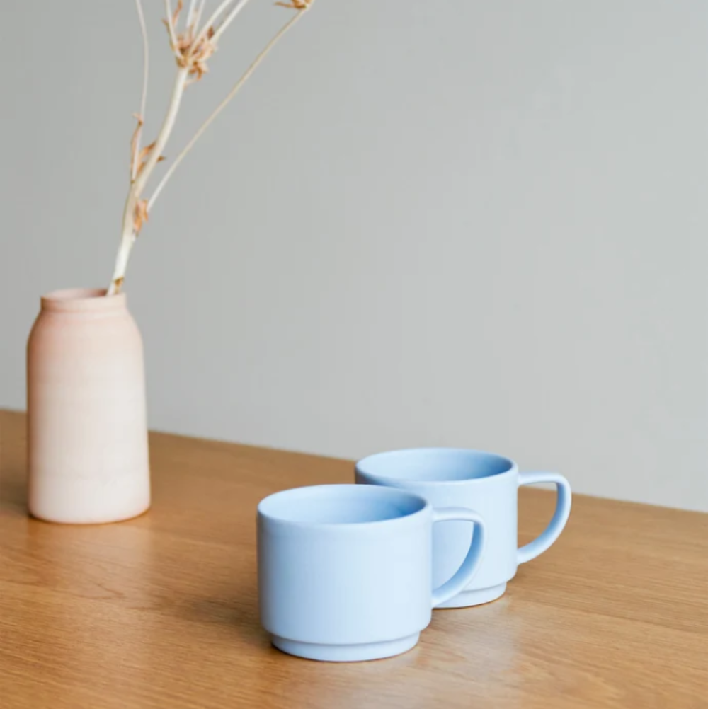 Two light blue ceramic mugs on a wooden table with a minimalist pink vase and dried stems in the background.