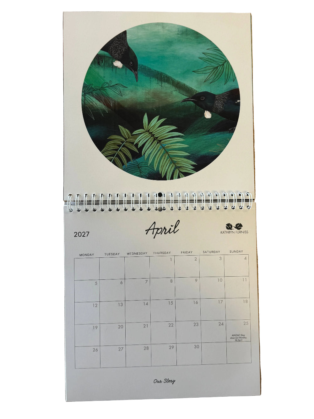 A spiral-bound desk calendar page from One Shop for 2027, featuring a circular illustration of two penguins in a green tropical scene above a monthly calendar grid, photographed on a wooden surface.