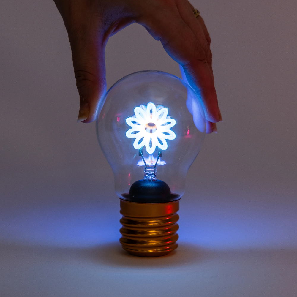 A hand is holding a clear glass light bulb with a glowing white flower-shaped bulb inside and a gold screw base, set against a dark gray background.
