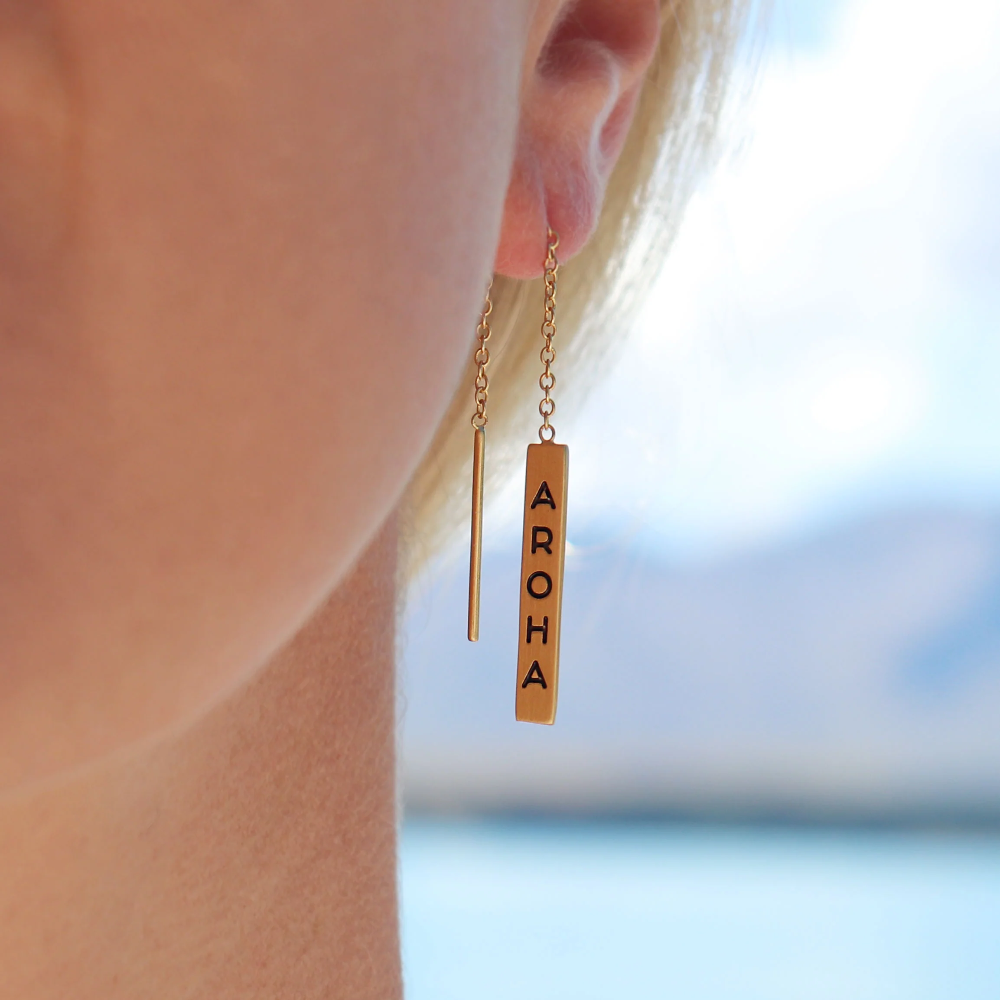 A close-up of a person's ear wearing a gold drop earring with a vertical bar engraved with the word 'AROA'.