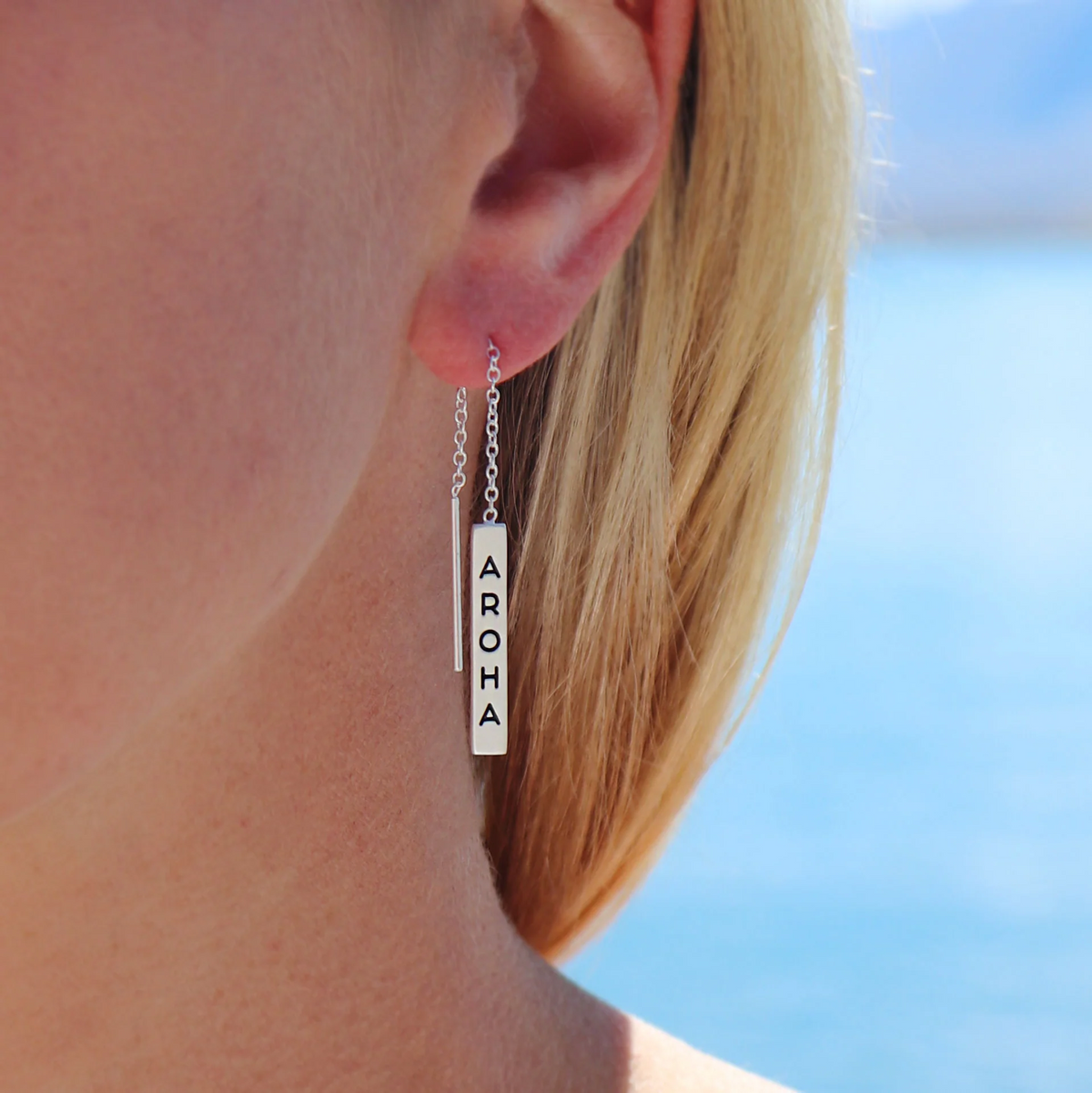 Silver earring with 'AROHA' pendant worn by a person against a blue sky background