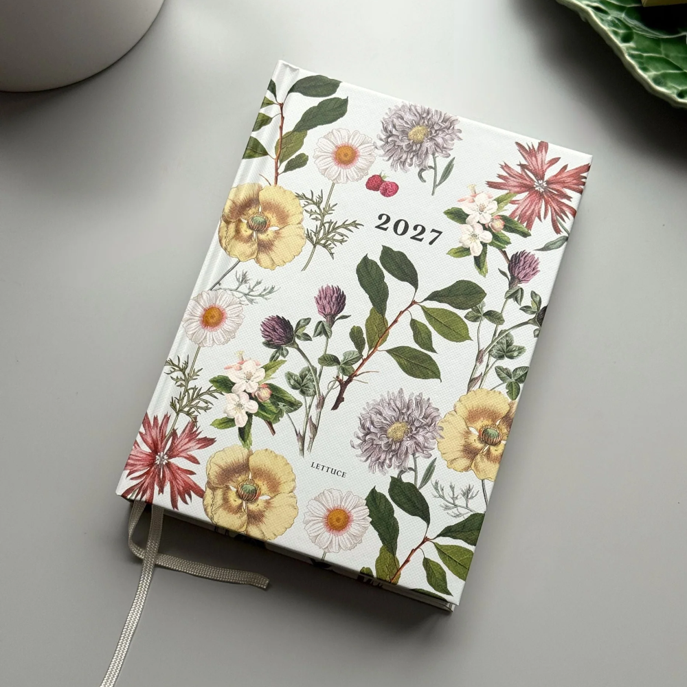 A 2027 floral planner or journal with a white cover decorated with colorful flowers and green leaves, shown on a gray tabletop with a ribbon bookmark.