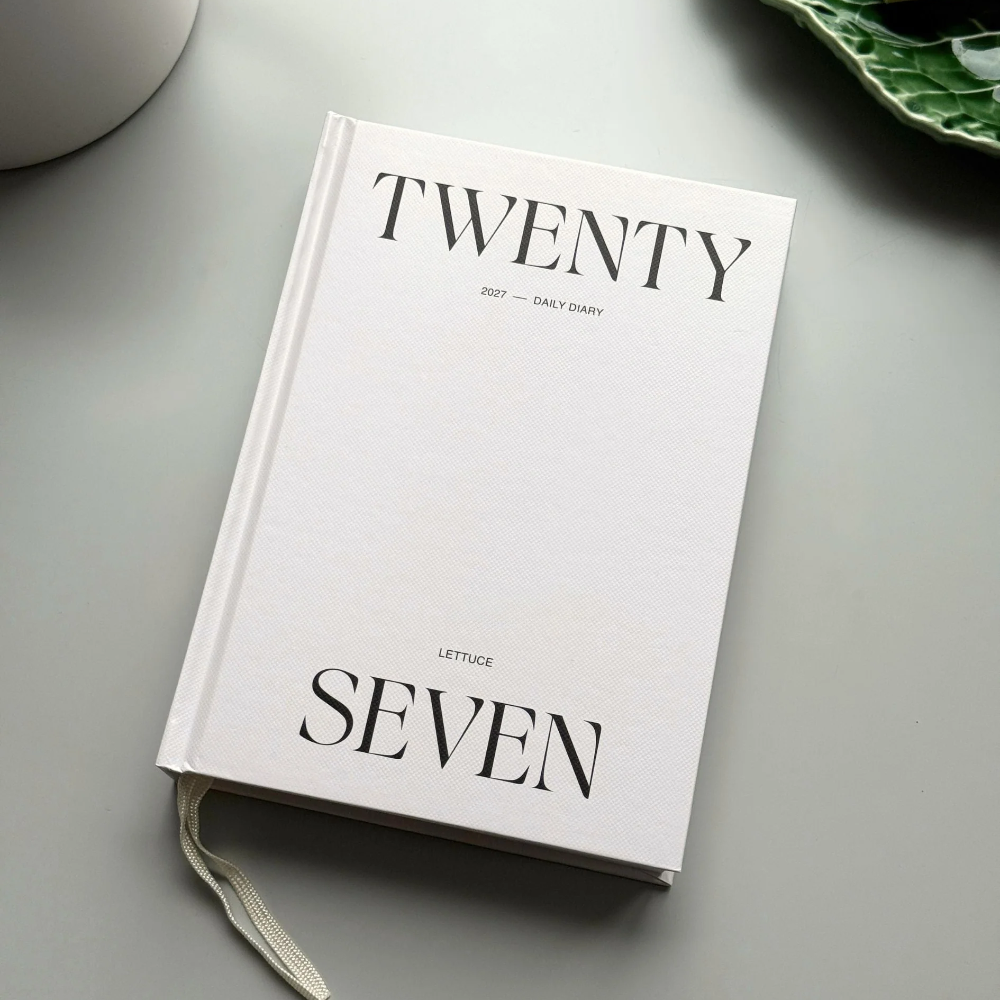 A white hardcover daily journal titled 'TWENTY SEVEN' by Lettrice, styled on a light gray surface with a white ceramic mug and a green leaf-patterned plate in the background.