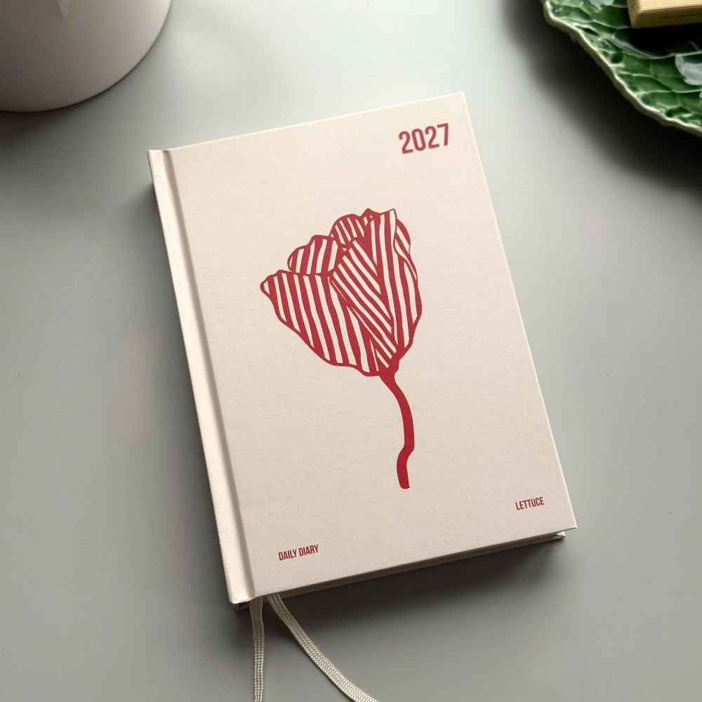 A 2027 daily letter planner with a cream-colored cover, featuring a red striped tulip illustration and a white ribbon bookmark, styled on a gray surface with a white teacup and a green leaf-shaped dish in the background.