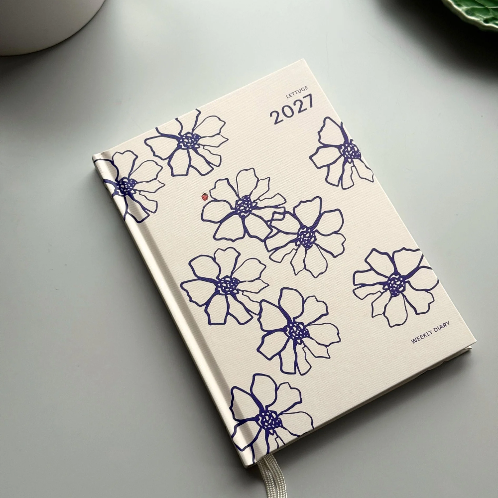 A cream-colored 2027 daily planner with blue floral line drawings, shown on a gray surface with a white ceramic cup and a green leaf-shaped dish partially visible.