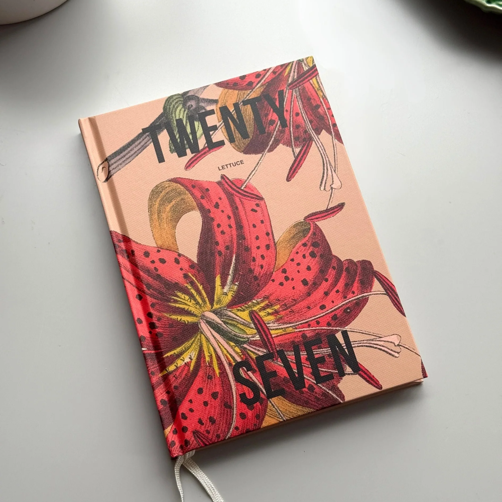 A hardcover notebook titled 'Twenty Seven' with a peach cover and a large red lily floral illustration, photographed on a light gray surface with partial props visible at the edges.