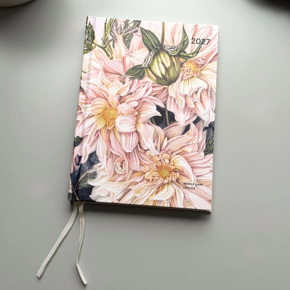 A hardcover planner or journal with a floral cover design featuring pink dahlia flowers and green leaves, shown on a light gray surface with a white ribbon bookmark.