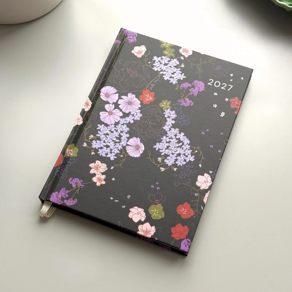 A 2027 daily planner with a black cover decorated with a colorful floral pattern, resting on a light gray surface with a white ceramic mug partially visible in the top left.