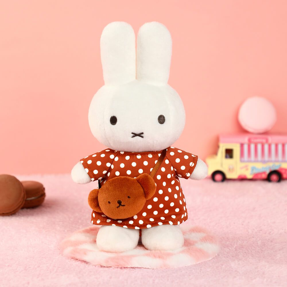 A white plush bunny doll wearing a rust-orange dress with white polka dots and carrying a small brown teddy bear purse, standing on a pink-and-white striped rug against a soft pink background with toy props.