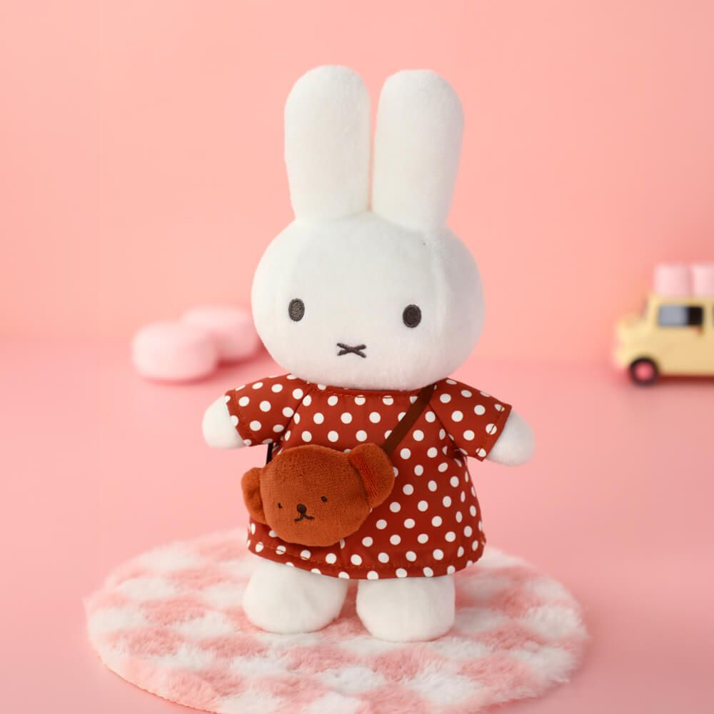 White plush rabbit toy with a red polka dot dress and brown bag on a pink background
