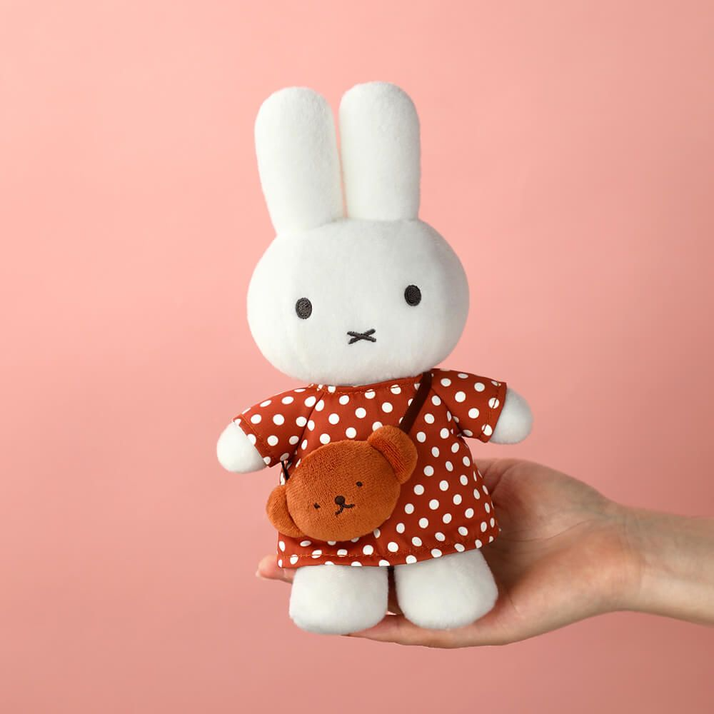 White plush rabbit toy with a red polka dot dress held against a pink background