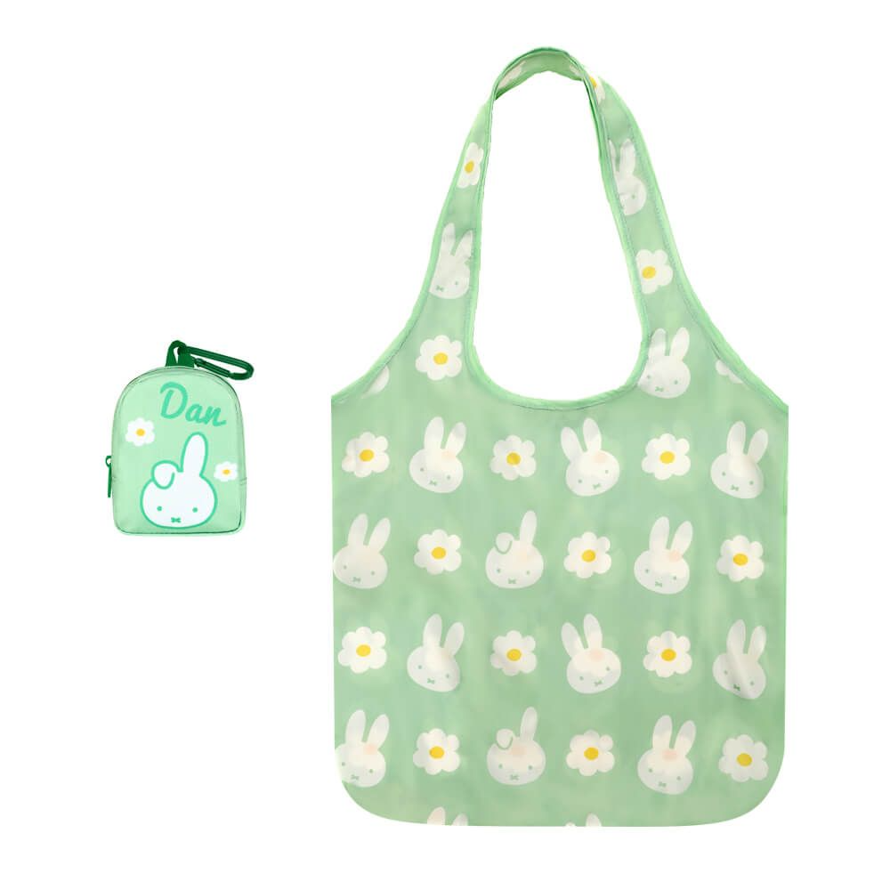 A light green reusable tote bag with a repeating pattern of white bunnies and daisies, shown alongside a matching small zip pouch with a green clip. The pouch has the name 'Dan' printed on it in white.