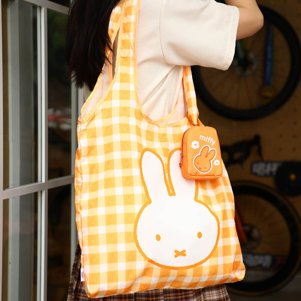 A person wearing a cream ribbed top carries a yellow and white gingham reusable tote bag with a large white bunny face graphic. A small matching orange pouch with a bunny graphic and the text 'miffy' is attached to the bag.