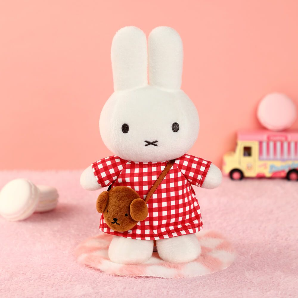 White plush rabbit toy in a red checkered dress with a brown bag on a pink background